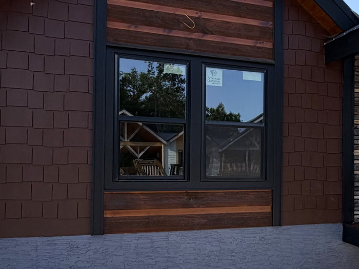 Finished elevation in cedar shake with a stained board band, black-framed window and standing-seam roof.